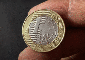 Moeda de 1 Real com a data 1998 sendo inspecionada de perto por uma pessoa. A imagem destaca a moeda rara que pode valer até R$ 3.000 devido à inscrição da letra 'P' de ensaio monetário.