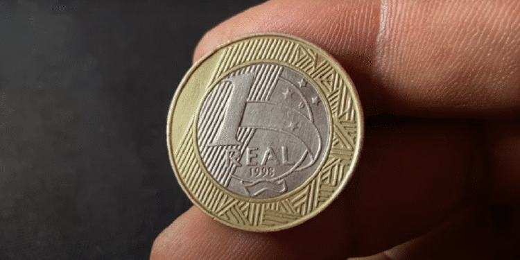 Moeda de 1 Real com a data 1998 sendo inspecionada de perto por uma pessoa. A imagem destaca a moeda rara que pode valer até R$ 3.000 devido à inscrição da letra 'P' de ensaio monetário.