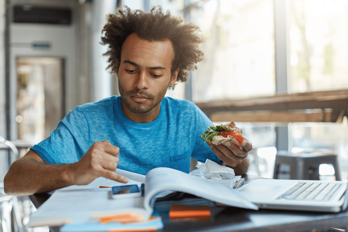 Prova Enem 2025 Homem jovem com camiseta azul estudando com livros e laptop enquanto come sanduíche na cafeteria iluminada