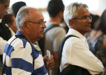 Dois homens idosos com óculos aguardando em uma fila com pessoas ao fundo desfocadas