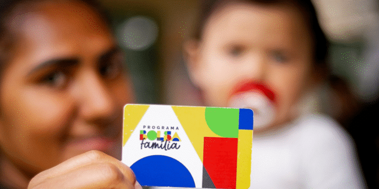Mulher segura o cartão do Programa Bolsa Família com um bebê ao fundo.