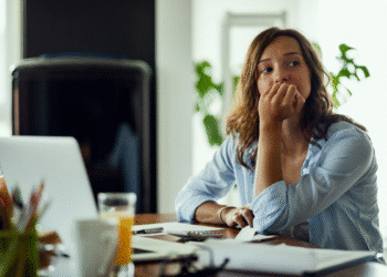 Mulher jovem pensativa sentada em mesa com laptop, documentos e copo de suco