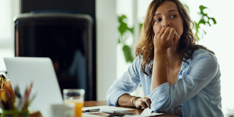 Mulher jovem pensativa sentada em mesa com laptop, documentos e copo de suco