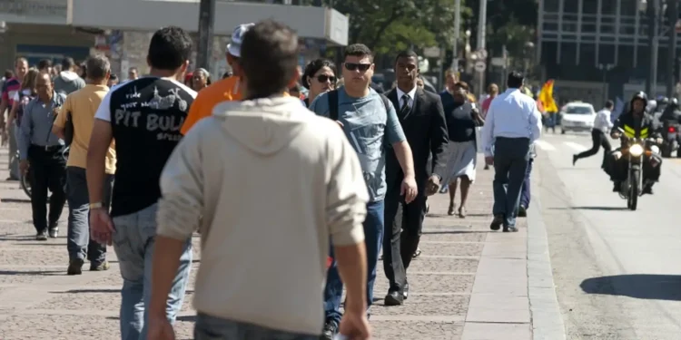 Pessoas diversas caminhando em uma calçada larga próxima à rua movimentada com veículos e motociclistas ao fundo
