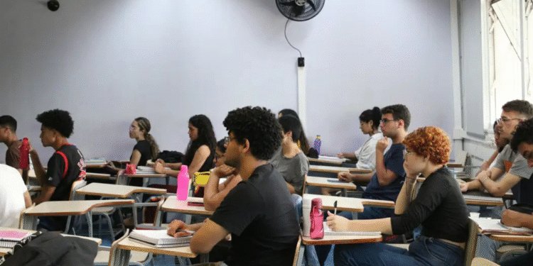 Sala de aula com diversos estudantes sentados em carteiras prestando atenção e tomando notas.