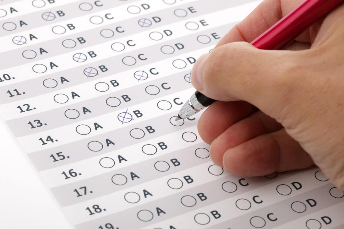 Mão segurando caneta preenchendo resposta em teste de múltipla escolha com opções A, B, C, D.