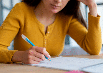 Mulher com blusa amarela escrevendo notas em um caderno aberto com caneta azul.