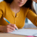 Mulher com blusa amarela escrevendo notas em um caderno aberto com caneta azul.