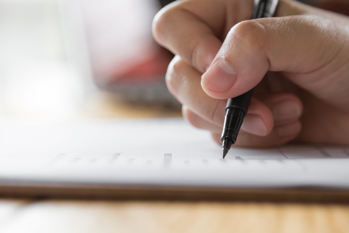 Concursos Câmara Municipal Foto da mão segurando caneta preta escrevendo em folha branca sobre superfície de madeira