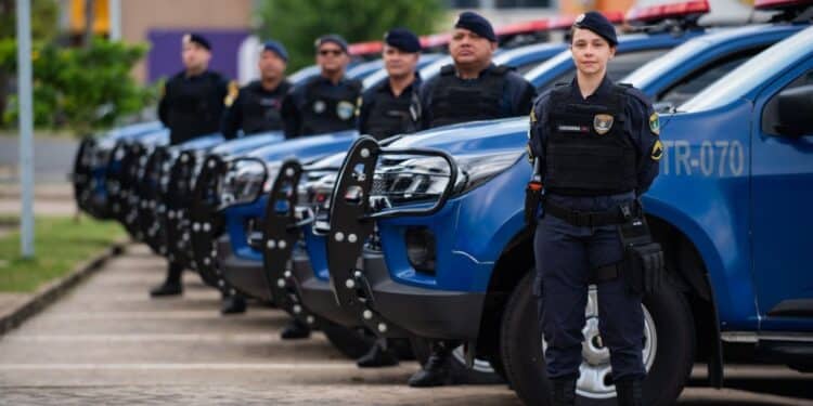 Guardas Municipais em formação alinhada ao lado de várias viaturas policiais azuis em ambiente externo.