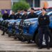 Grupo de Guardas Municipais em uniformes alinhados ao lado de viaturas azuis estacionadas