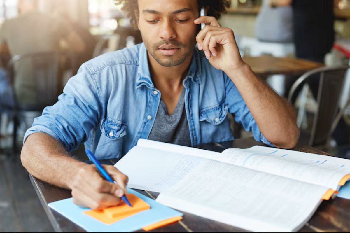 Dicas para se Destacar nos Concursos da Guarda Municipal Jovem estudando e fazendo anotações enquanto conversa ao telefone, em um ambiente descontraído, com livros e material de estudo.