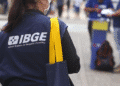 Funcionária do IBGE vista de costas usando colete azul com logo e máscara, carregando bolsa azul com alça amarela.