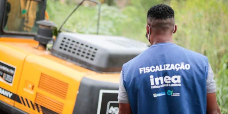 Fiscal do INEA usando colete azul de fiscalização ambiental próximo a máquina amarela em área verde