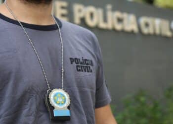 Homem com camisa cinza da Polícia Civil exibindo distintivo em corrente no peito em frente à delegacia.
