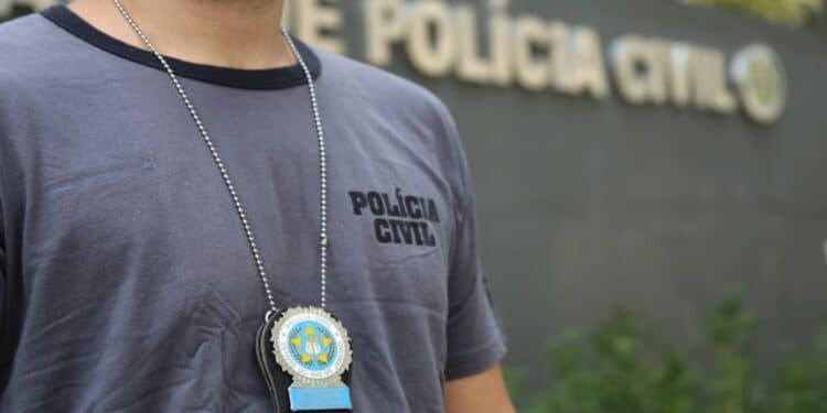 Homem com camisa cinza da Polícia Civil exibindo distintivo em corrente no peito em frente à delegacia.