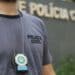 Homem com camisa cinza da Polícia Civil exibindo distintivo em corrente no peito em frente à delegacia.