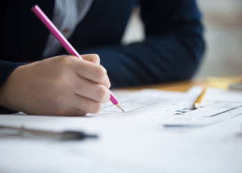 Mão segurando lápis cor de rosa desenhando plantas arquitetônicas sobre mesa com ferramentas de desenho