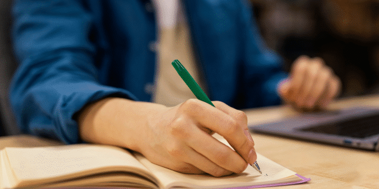 Mão segurando caneta verde escrevendo em caderno aberto com pessoa ao fundo e notebook ao lado