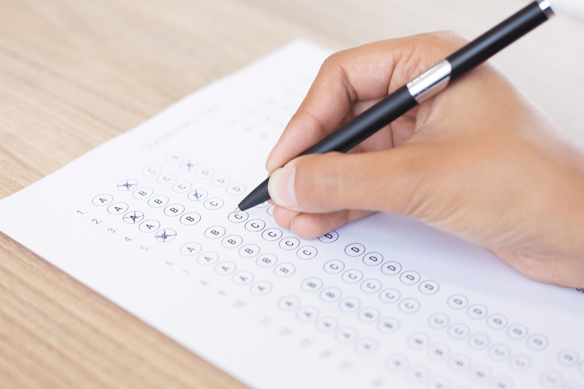 Mão preenchendo respostas de prova objetiva com caneta preta em folha branca sob superfície de madeira.