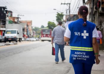 Mulher vestindo colete azul do SUS, agente comunitário de saúde, andando por rua urbana movimentada