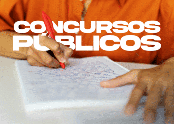 Pessoa escrevendo com caneta vermelha em caderno durante estudo para concursos públicos, vestindo camisa laranja.
