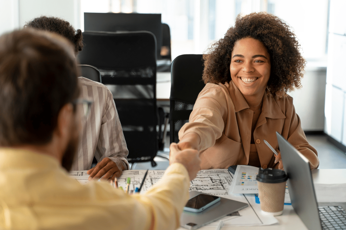 Duas pessoas apertando as mãos em ambiente de trabalho com laptop, café e documentos na mesa.