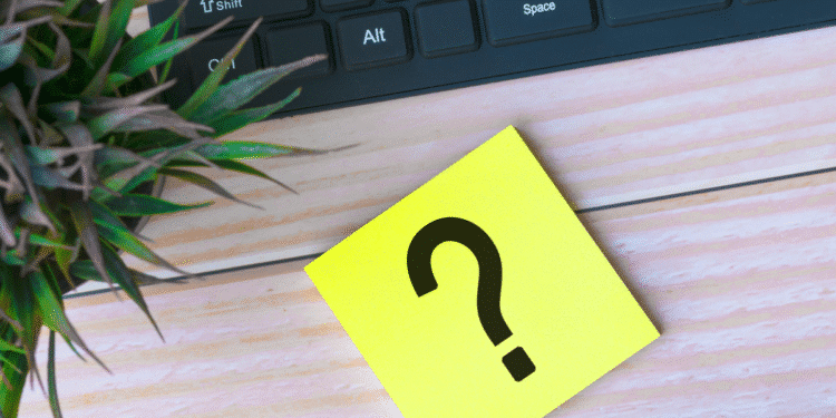 Post-it amarelo com ponto de interrogação preto ao lado de teclado e planta sobre mesa de madeira clara
