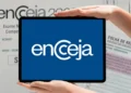 Logo do Encceja em fundo azul exibido em tablet segurado por mãos, com documentos de exame ao fundo.