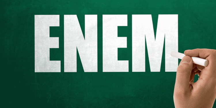 Mão segurando giz em frente a quadro verde com a palavra ENEM escrita em destaque.