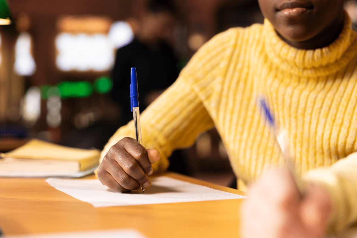 Mão de estudante com blusa amarela escrevendo em folha de papel com caneta azul em mesa de madeira