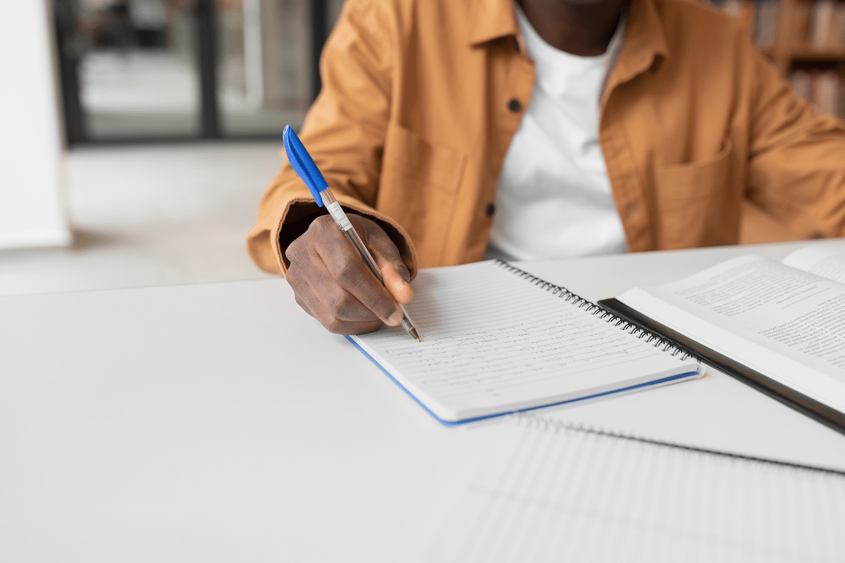 Pessoa com camisa marrom escrevendo anotações em caderno espiral com caneta azul em mesa branca.