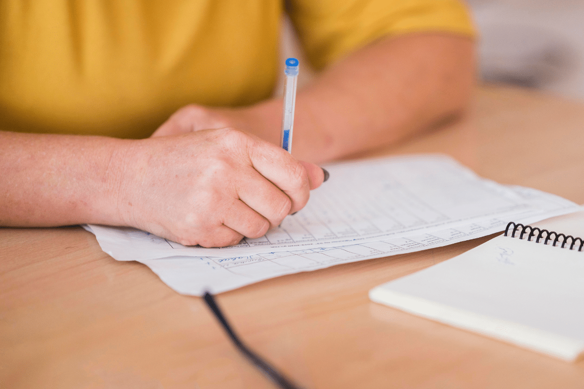 Mão segurando caneta azul escrevendo em folhas de papel sobre mesa de madeira clara