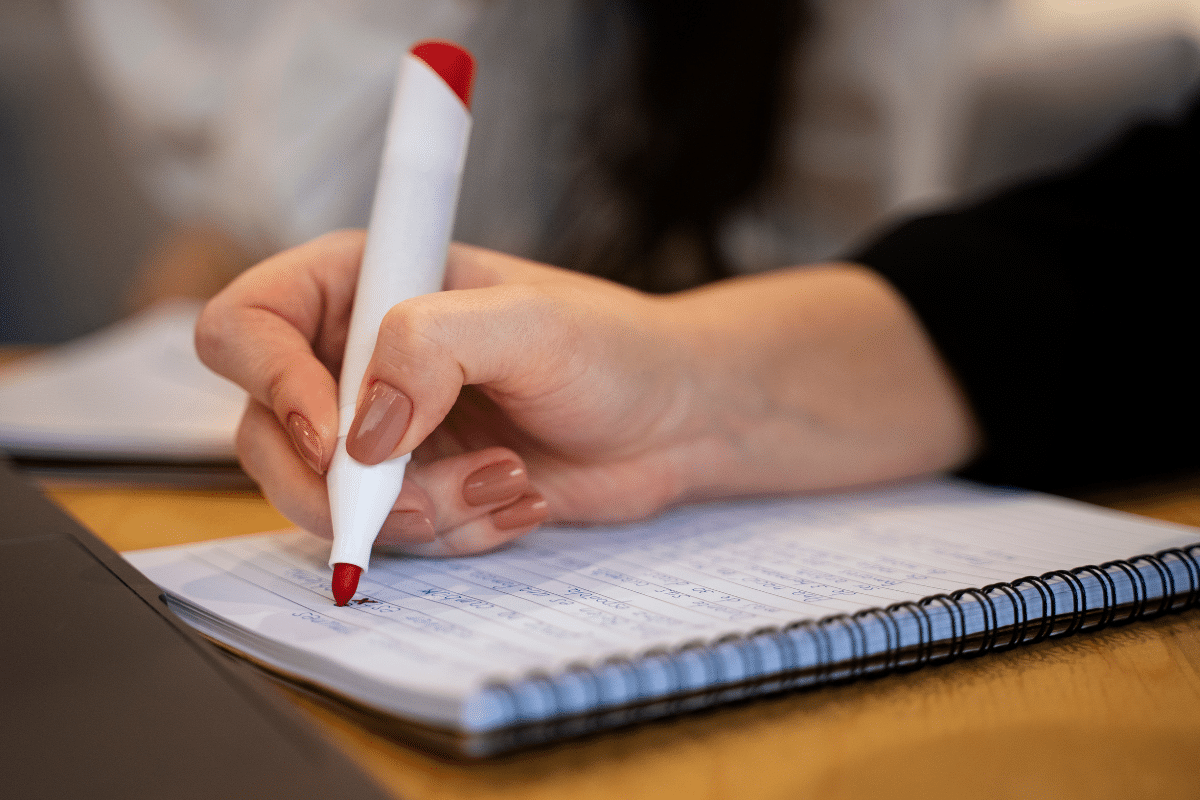 Mão com unhas pintadas segurando caneta vermelha escrevendo em caderno espiral com anotações à mão
