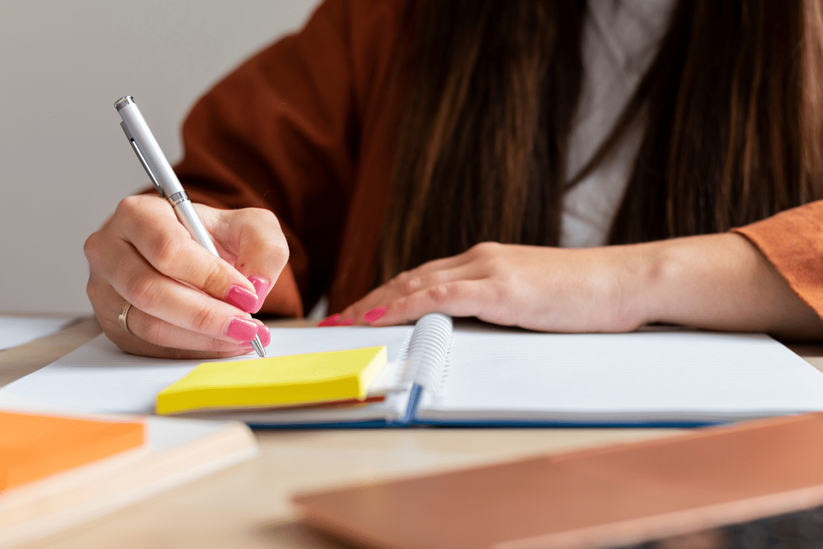 Mão feminina com unha rosa escrevendo em caderno espiral aberto com caneta prata e post-its coloridos na mesa