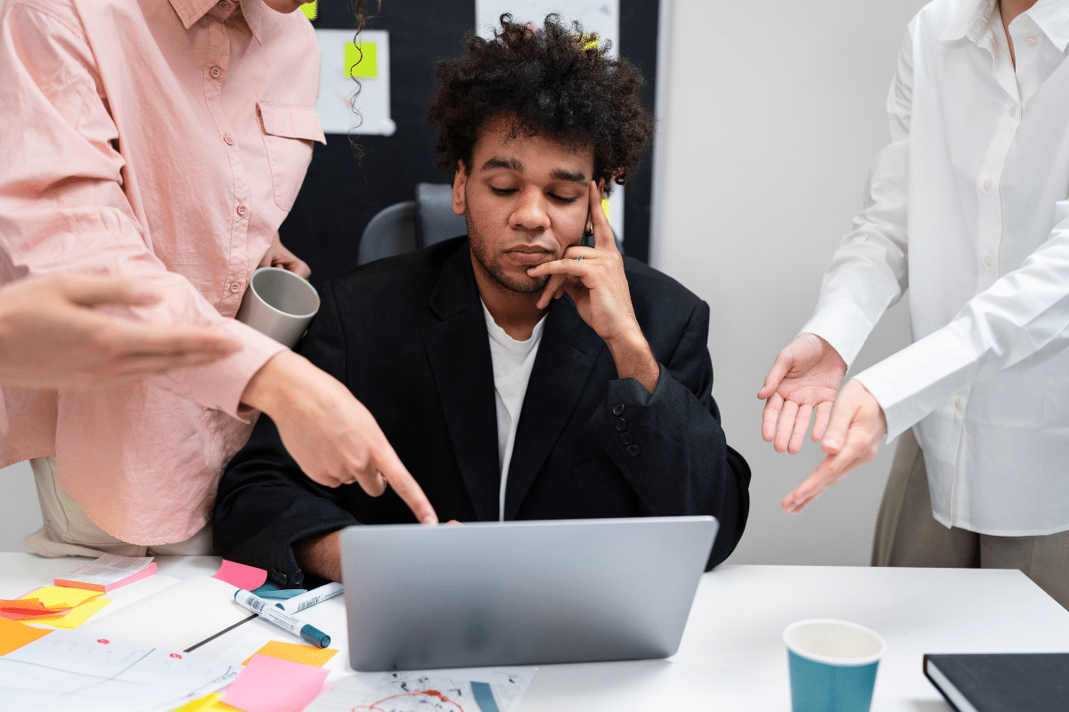 Homem pensativo e preocupado em frente a laptop enquanto colegas apontam e discutem