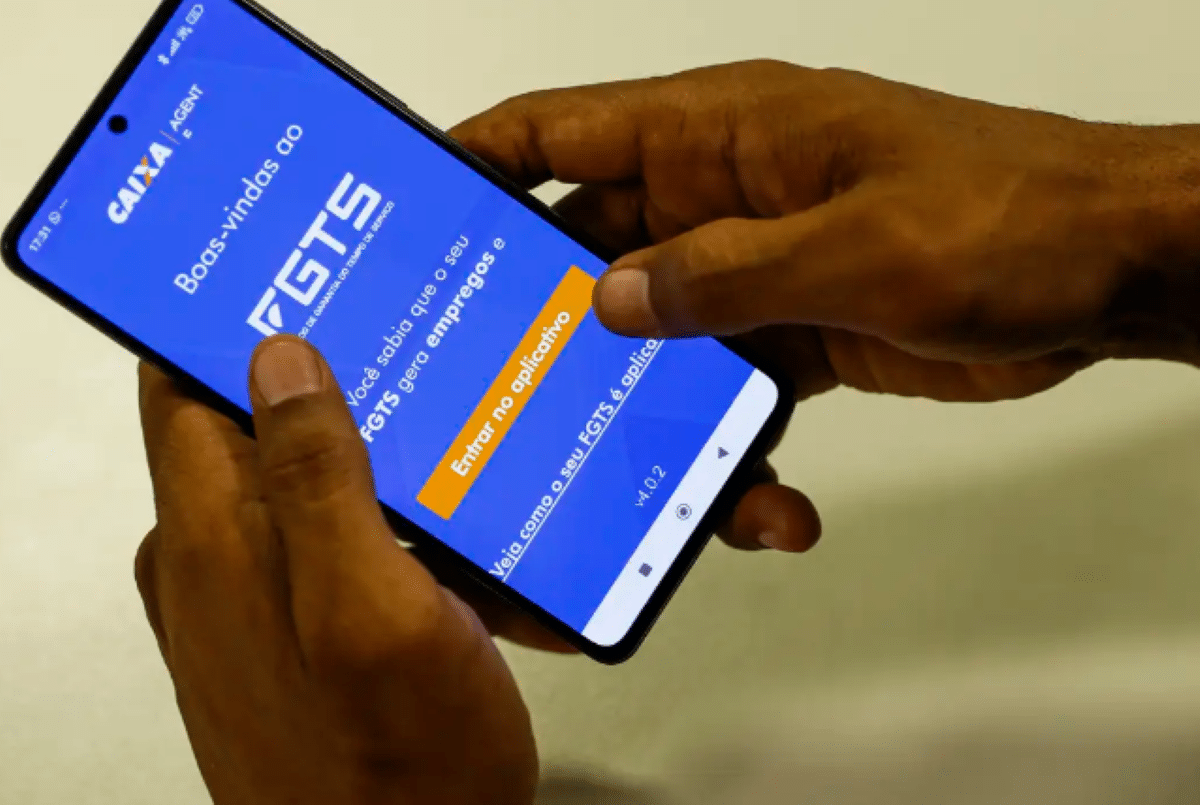 Saque-Aniversário do FGTS: Nascidos em Setembro, Outubro e Novembro podem receber; veja o prazo e novas regras Mãos segurando smartphone com tela inicial do aplicativo FGTS da Caixa, exibindo botão laranja "Entrar no aplicativo" e mensagem de boas-vindas.