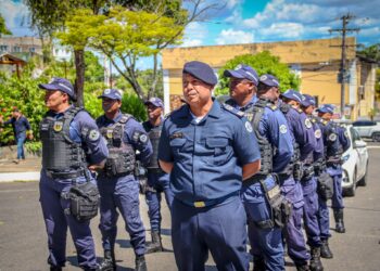 Grupo de guardas municipais uniformizados em formação na rua sob céu azul claro com árvores ao fundo