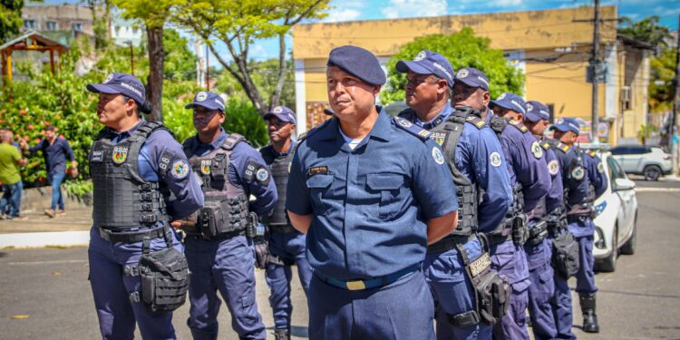 Grupo de guardas municipais uniformizados em formação na rua sob céu azul claro com árvores ao fundo