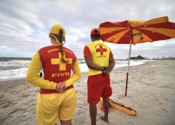 Dois guardas-vidas de costas na praia com uniformes vermelhos e amarelos observando o mar sob céu nublado