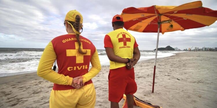 Dois guardas-vidas de costas na praia com uniformes vermelhos e amarelos observando o mar sob céu nublado