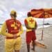 Dois guardas-vidas de costas na praia com uniformes vermelhos e amarelos observando o mar sob céu nublado