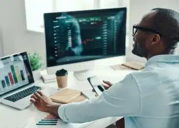 Homem sorridente e de óculos em ambiente de escritório analisando gráficos de mercado financeiro em seu monitor e notebook, representando a análise de tendências de carreira e salários para 2026.