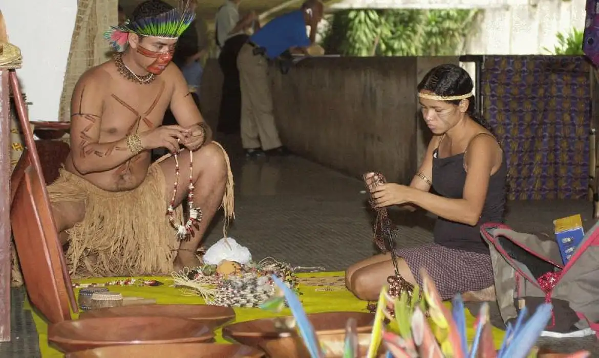 Artesanato indígena em ambiente cultural tradicional Homem e mulher indígenas confeccionando artesanato em ambiente fechado com vestimentas tradicionais