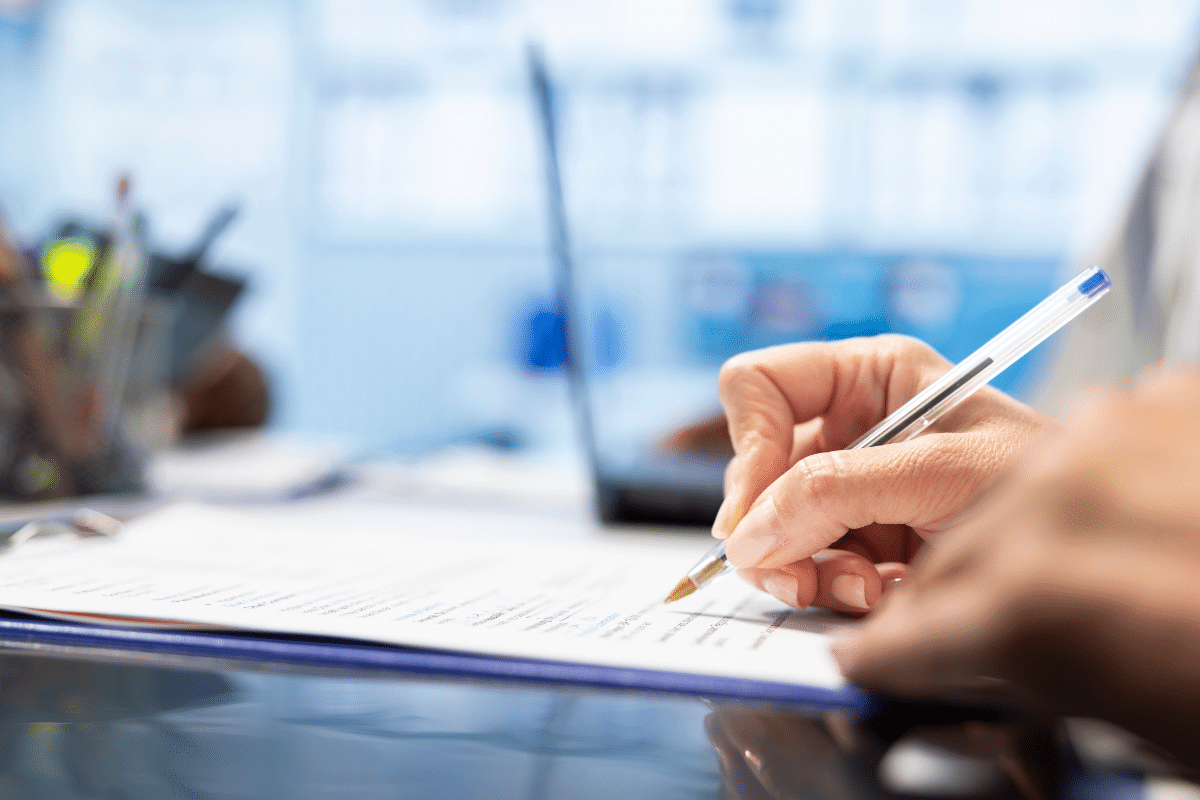 Mão escrevendo em documento usando caneta azul em ambiente de trabalho com laptop ao fundo