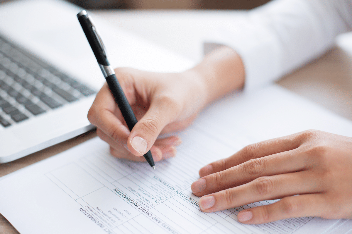 Inscrição Mão segurando caneta preenchendo formulário de informações comerciais ao lado de notebook