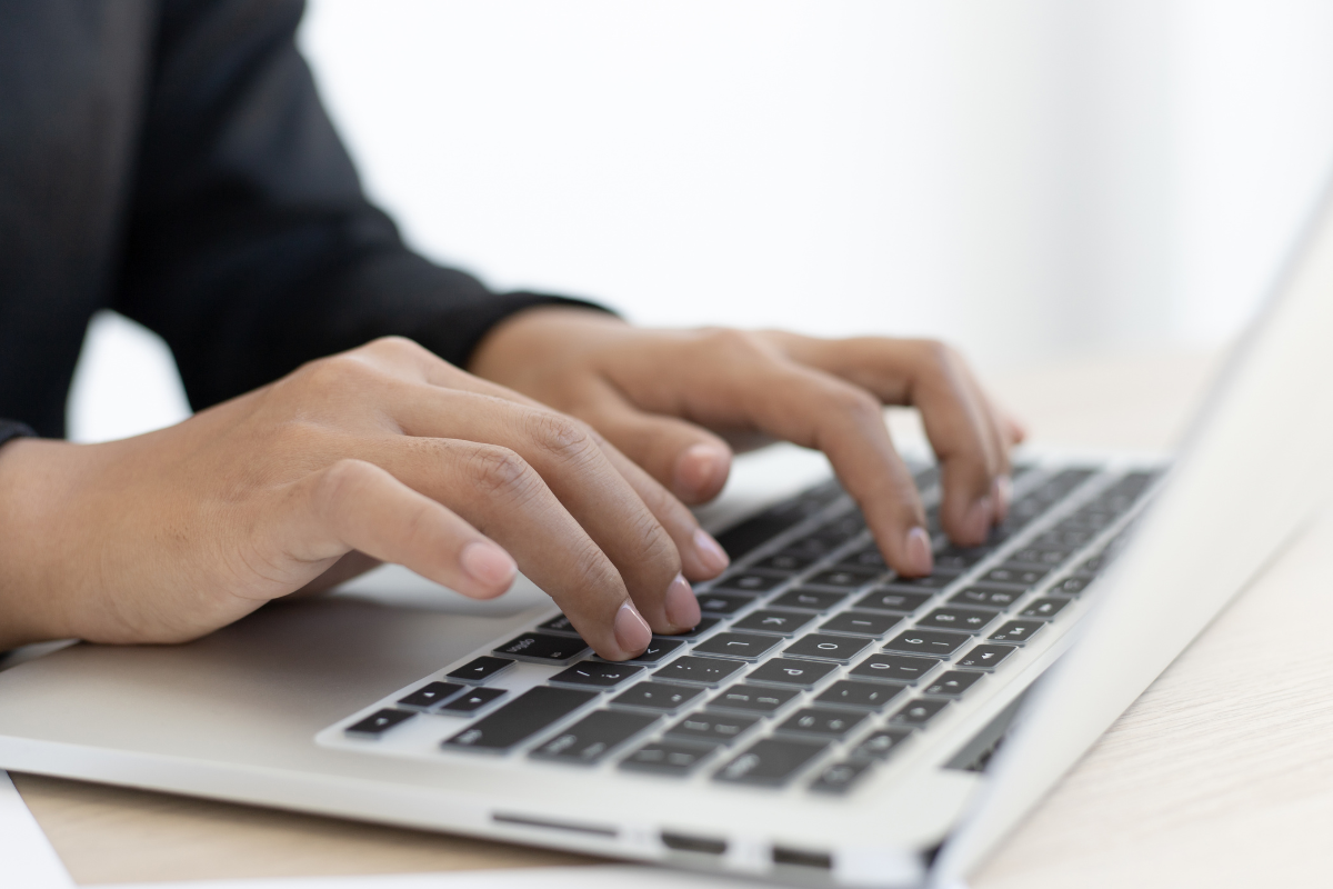 inscrição Foto das mãos de uma pessoa digitando em teclado de notebook prateado sobre mesa clara