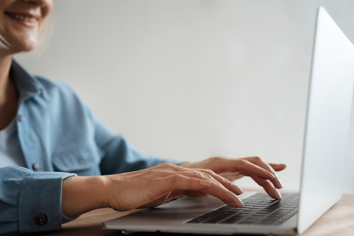 Mulher sorridente digitando no teclado de um notebook prateado em mesa de madeira clara