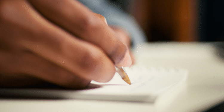 Imagem da mão escrevendo com caneta esferográfica em caderno branco sobre mesa clara