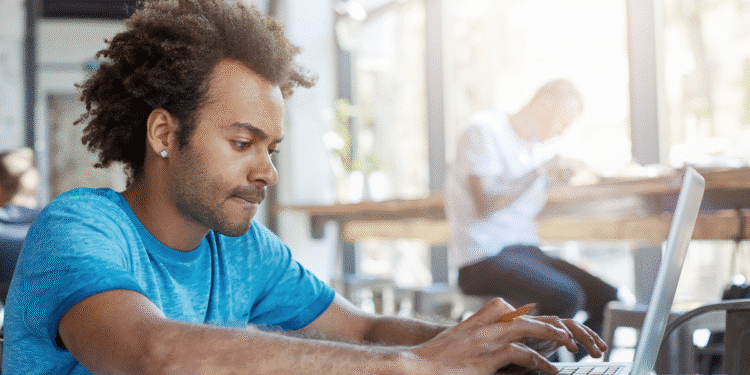 Homem jovem concentrado estudando no laptop em local de luz natural com livros e caderno à frente